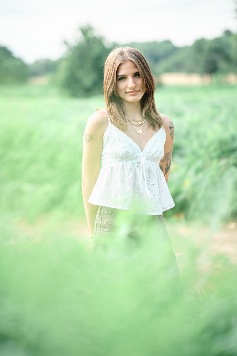Young woman in white lace top standing in lush green field, wearing layered gold necklaces and tattoo on arm.