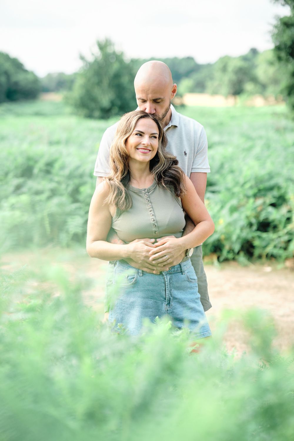 A couple embraces outdoors, man kissing woman's head while she smiles, surrounded by lush green nature.