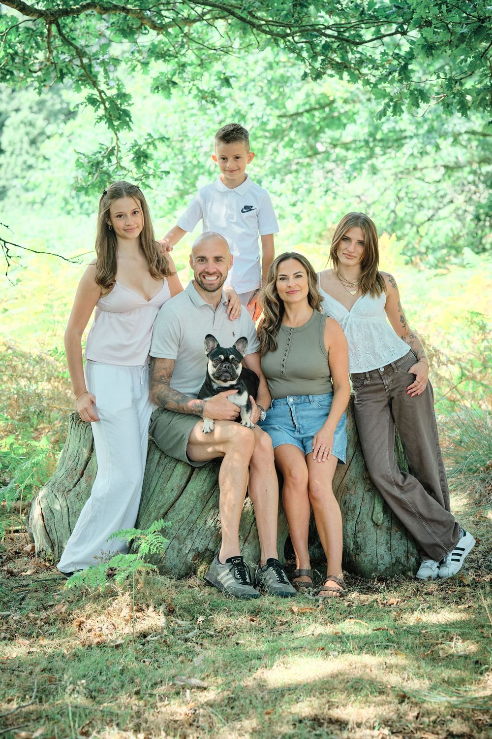 Happy family of four with tattoed dad holding French Bulldog, posing on tree stump in lush green forest.