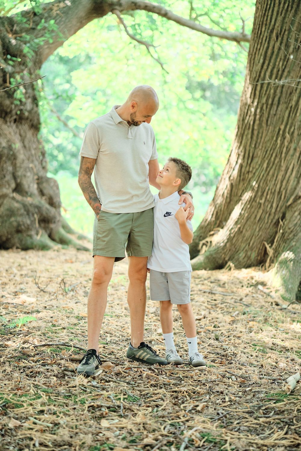 A bald father and young son share a tender moment in a sunlit forest, both wearing casual summer outfits.