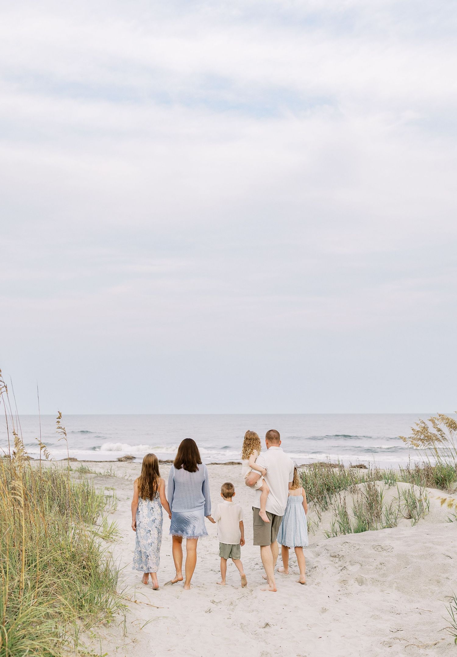 Family of five standing on sandy beach path facing ocean waves, dressed in coordinating blue and white outfits.