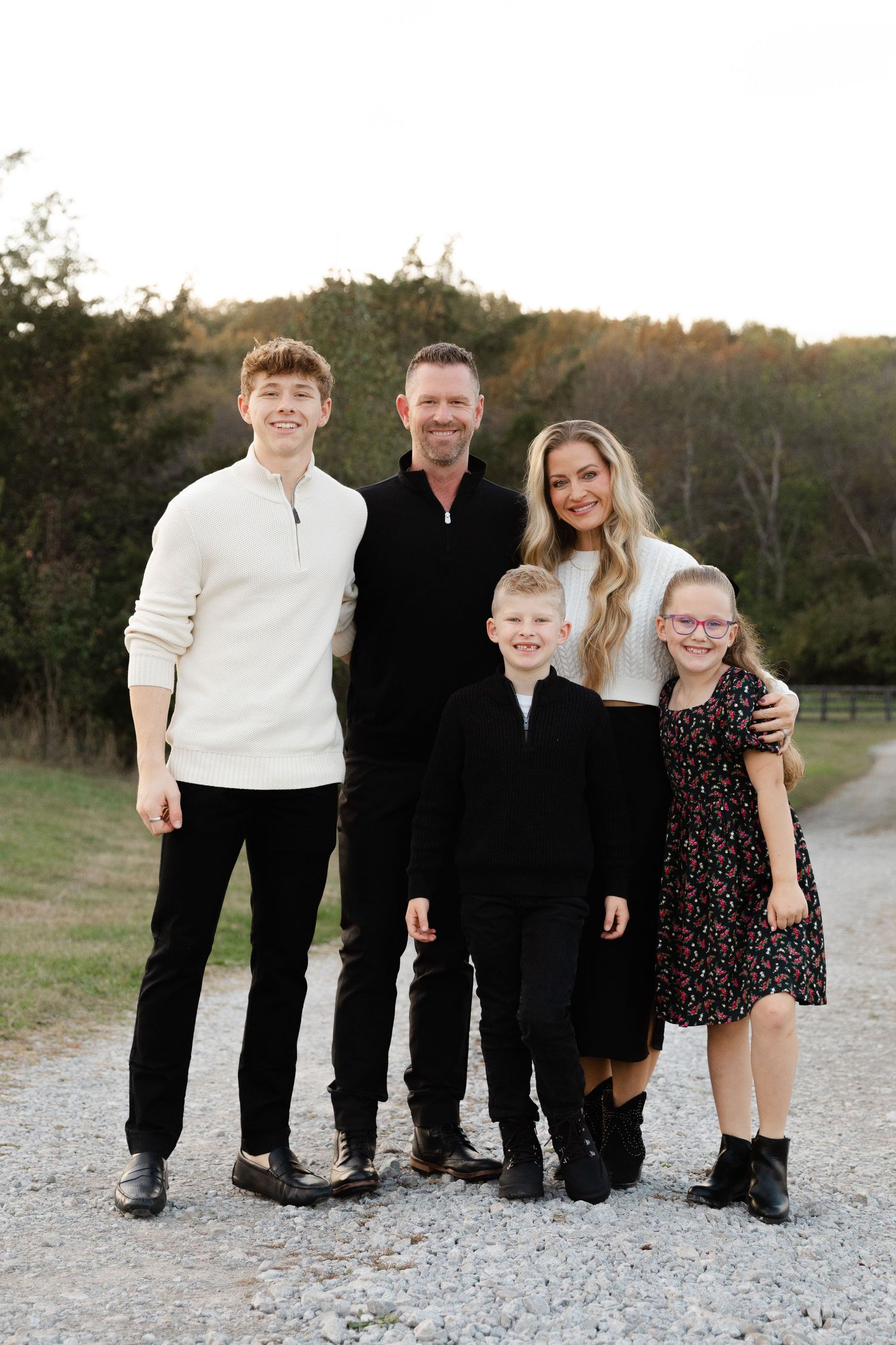 Family of five smiling together on a gravel path outdoors, dressed in black and white, with trees in the background.