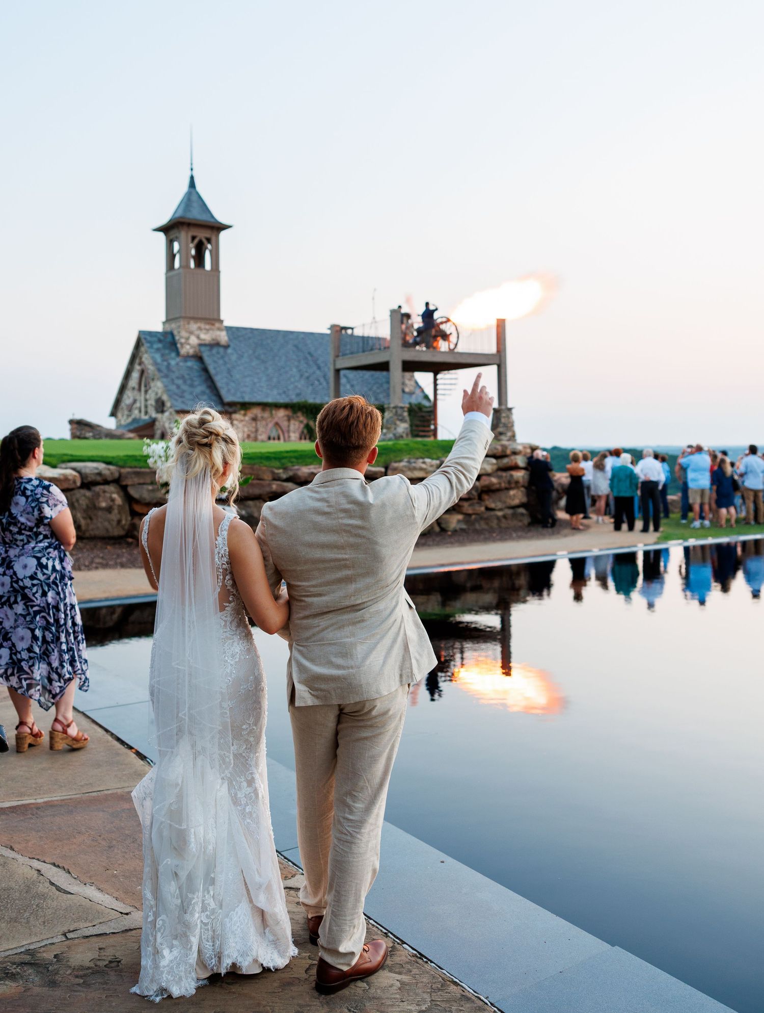 Bride and groom stand by reflective pool at dusk, pointing toward stone chapel with guests gathered outside.