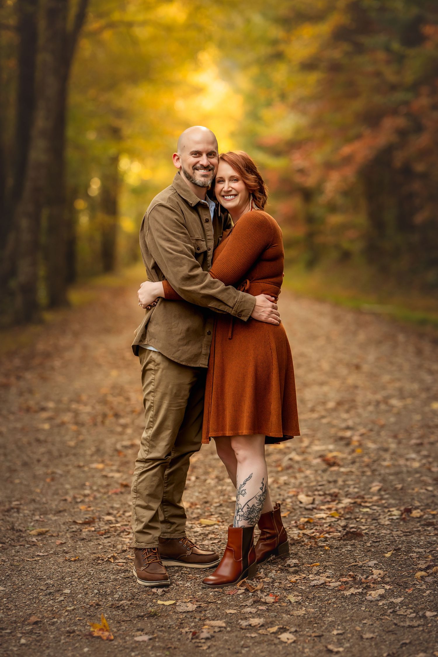 Couple embracing on a leaf-covered path surrounded by vibrant autumn foliage, woman in rust dress and man in olive jacket.
