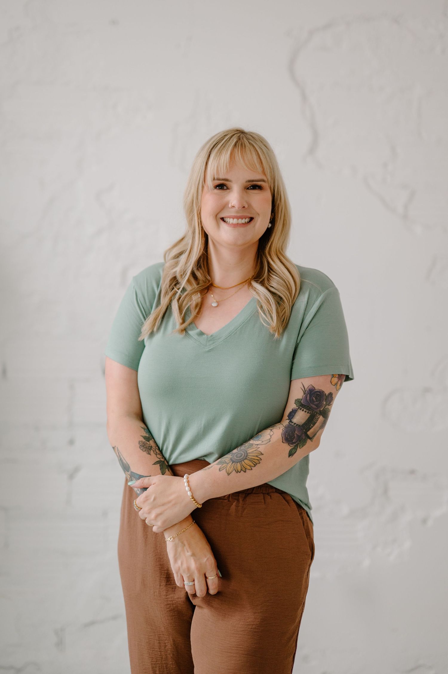 Smiling blonde woman with tattoos wearing a sage green top and brown pants poses against a white textured wall.