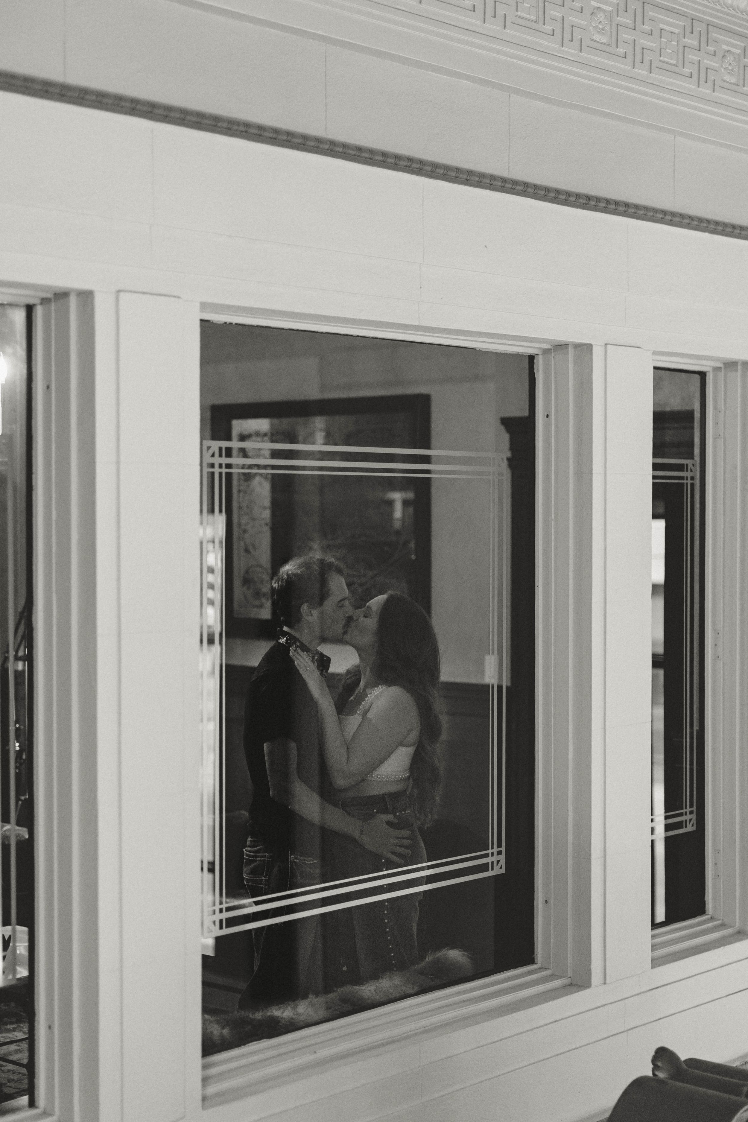 Black and white photo of couple kissing reflected through an ornate window with decorative glass.