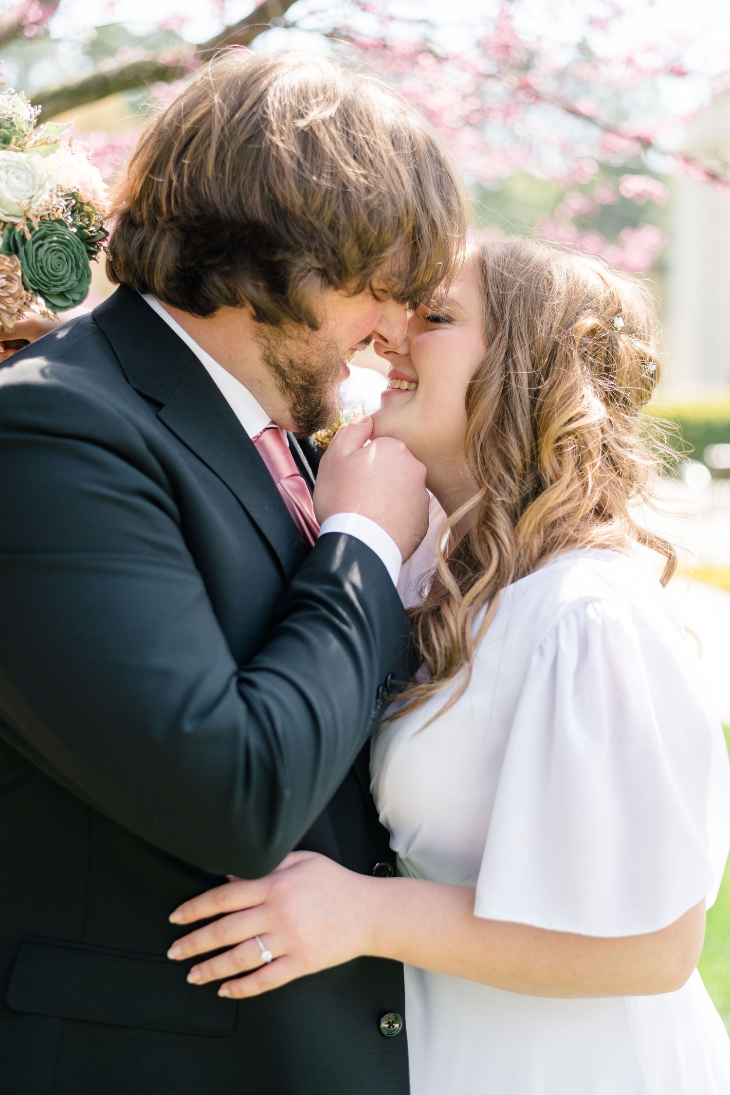 A bride and groom share a tender kiss outdoors near blooming cherry blossom trees on their wedding day.