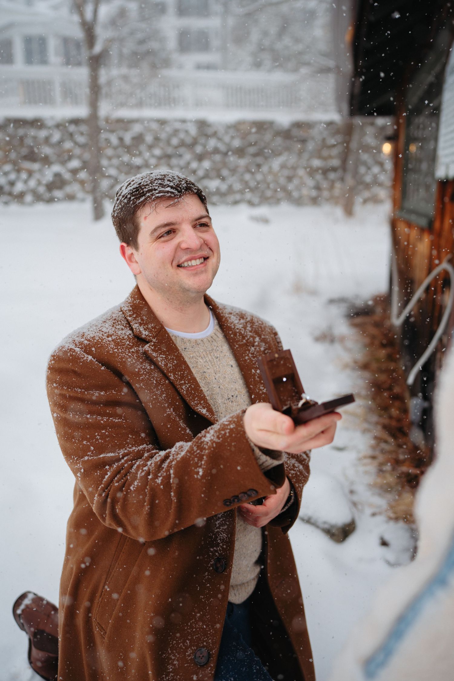 Smiling man in brown coat holding phone outdoors in snowfall, wearing sweater with snow dusting his shoulders.