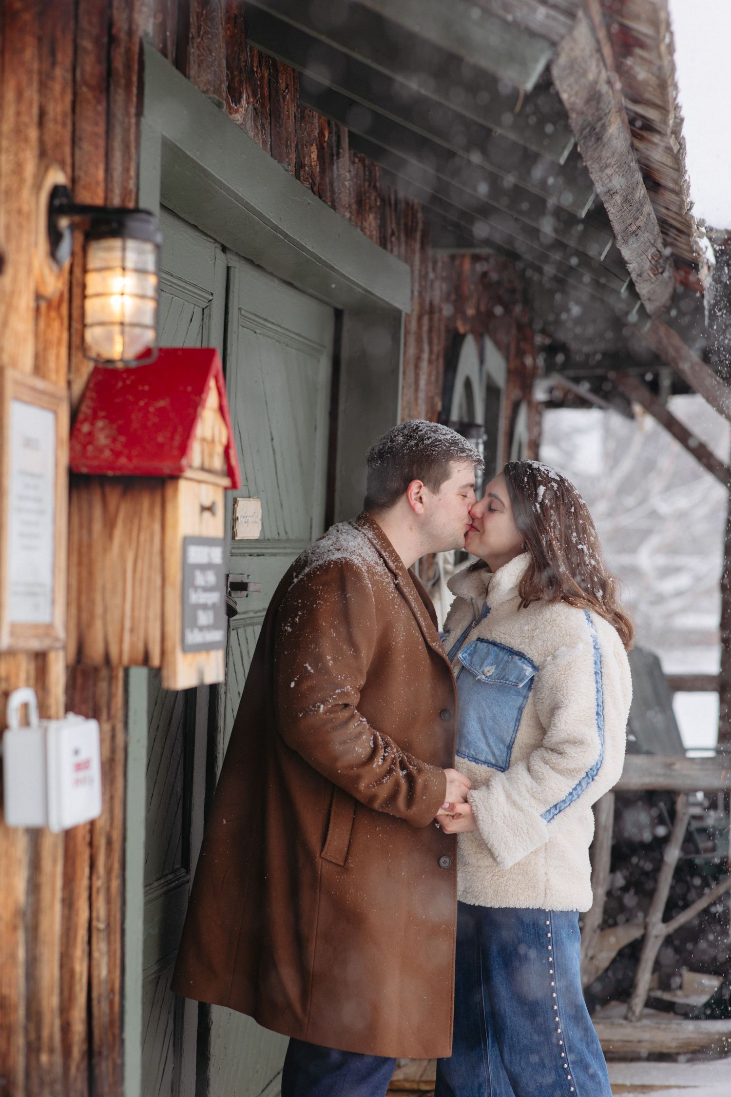 Couple kissing in the snow outside a rustic wooden cabin, wearing winter coats during a romantic snowy day.