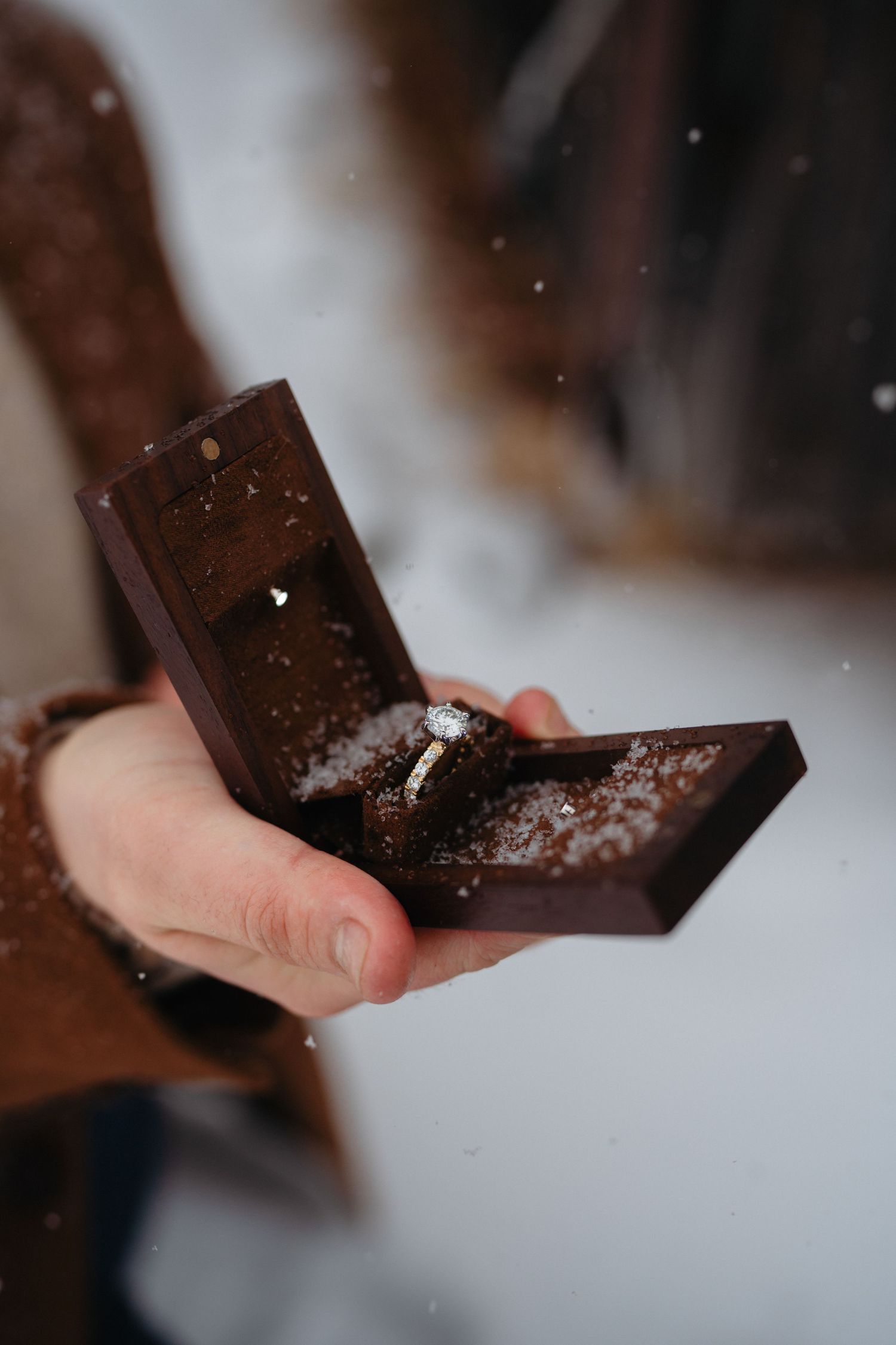 Hand holding a dark wooden ring box with a diamond engagement ring inside, with snow falling in the background.