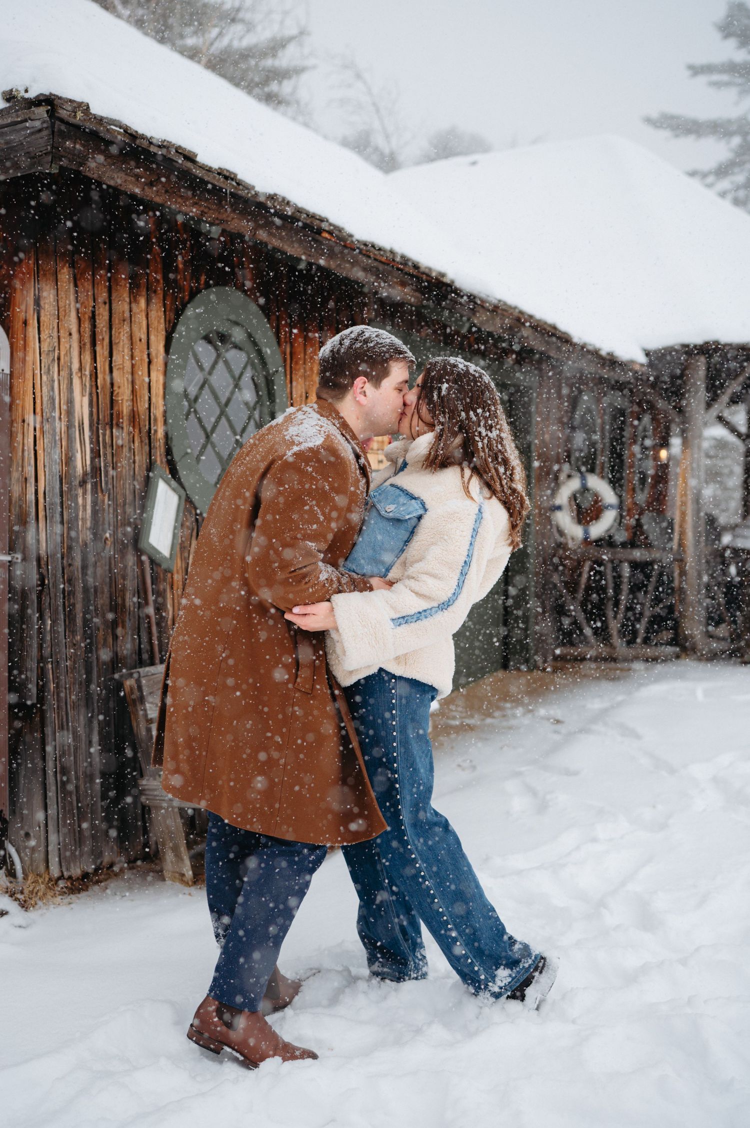 Couple sharing a romantic kiss in falling snow outside a rustic wooden cabin in winter.