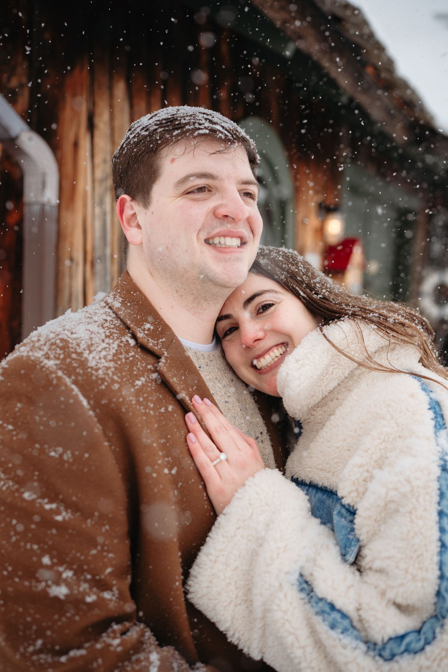 Smiling couple embracing in snowfall, woman in white fleece jacket leans on man in brown coat near rustic wooden building.