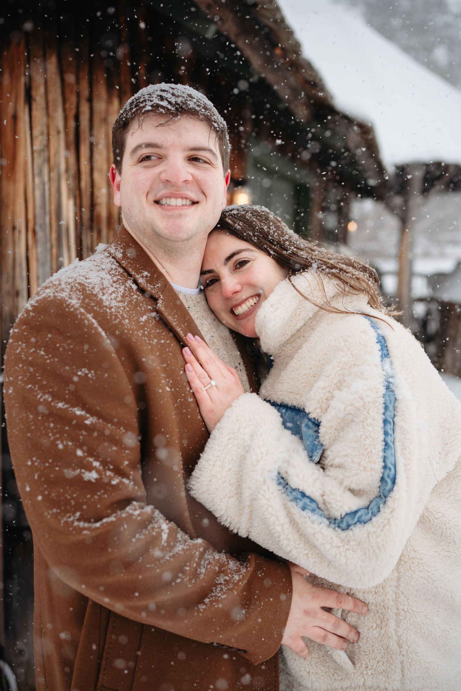 Smiling couple embracing in snowfall, man in brown coat and woman in white fleece jacket, posing outdoors near rustic building.