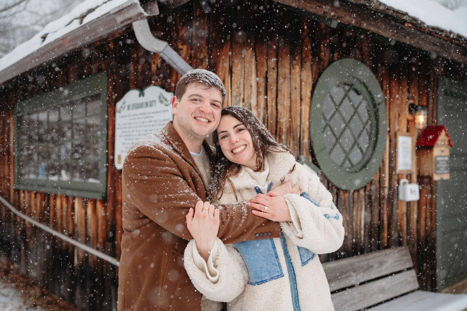 Happy couple embracing in snow outside rustic wooden cabin during winter snowfall.