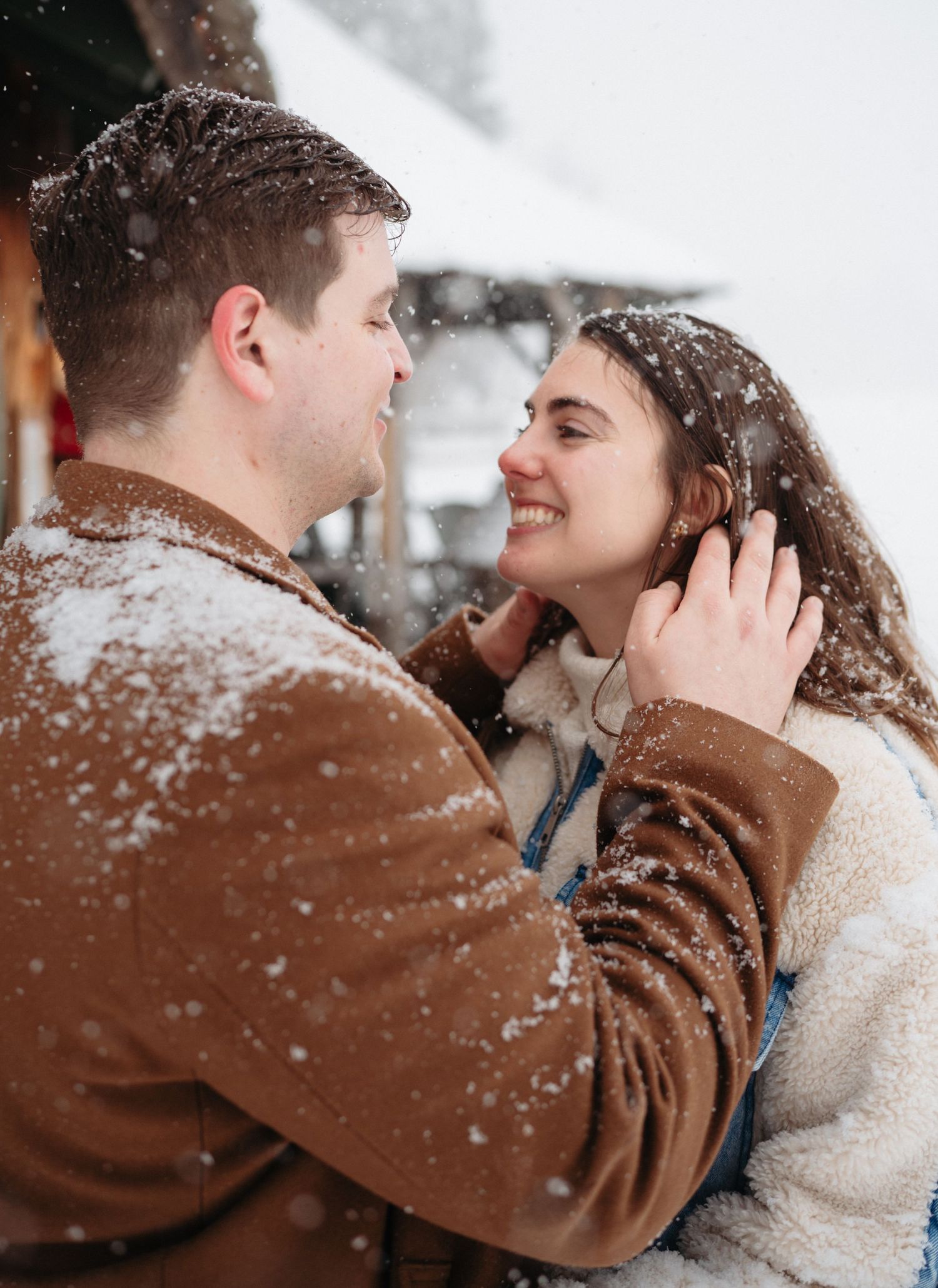 Couple embracing and smiling in falling snow, wearing cozy winter jackets outdoors.