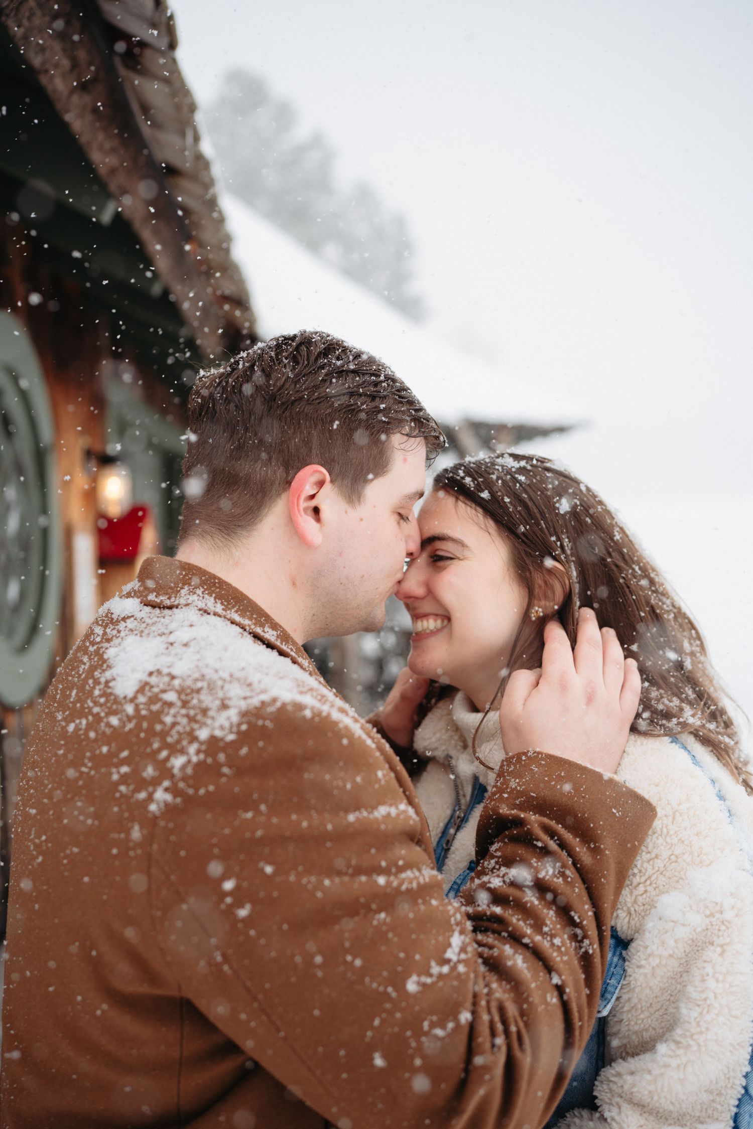 Romantic couple sharing a tender moment in snowfall, wearing winter coats, smiling face to face outdoors.