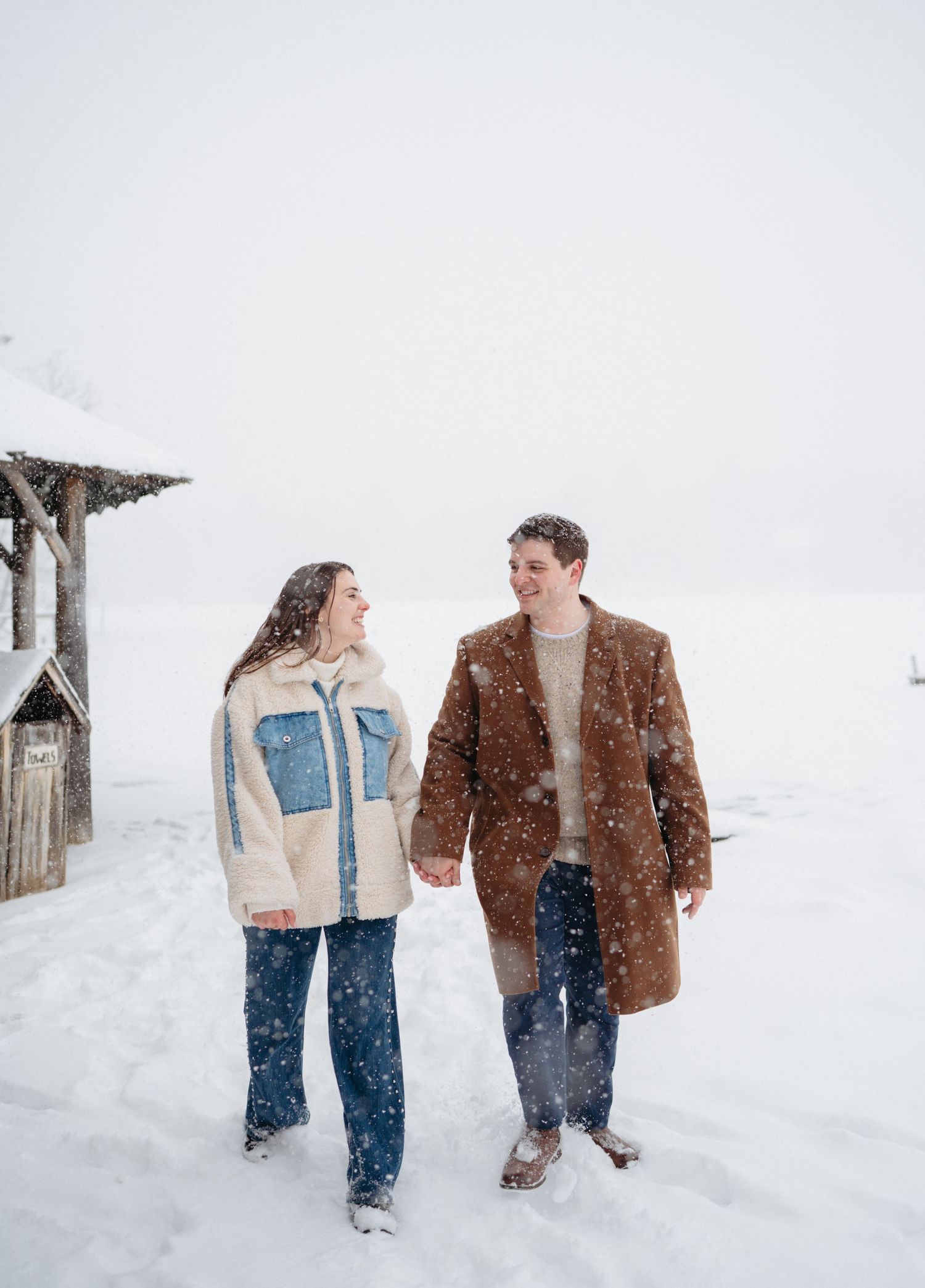 Couple holding hands and smiling while walking in snowfall, wearing winter coats in a snowy outdoor setting.