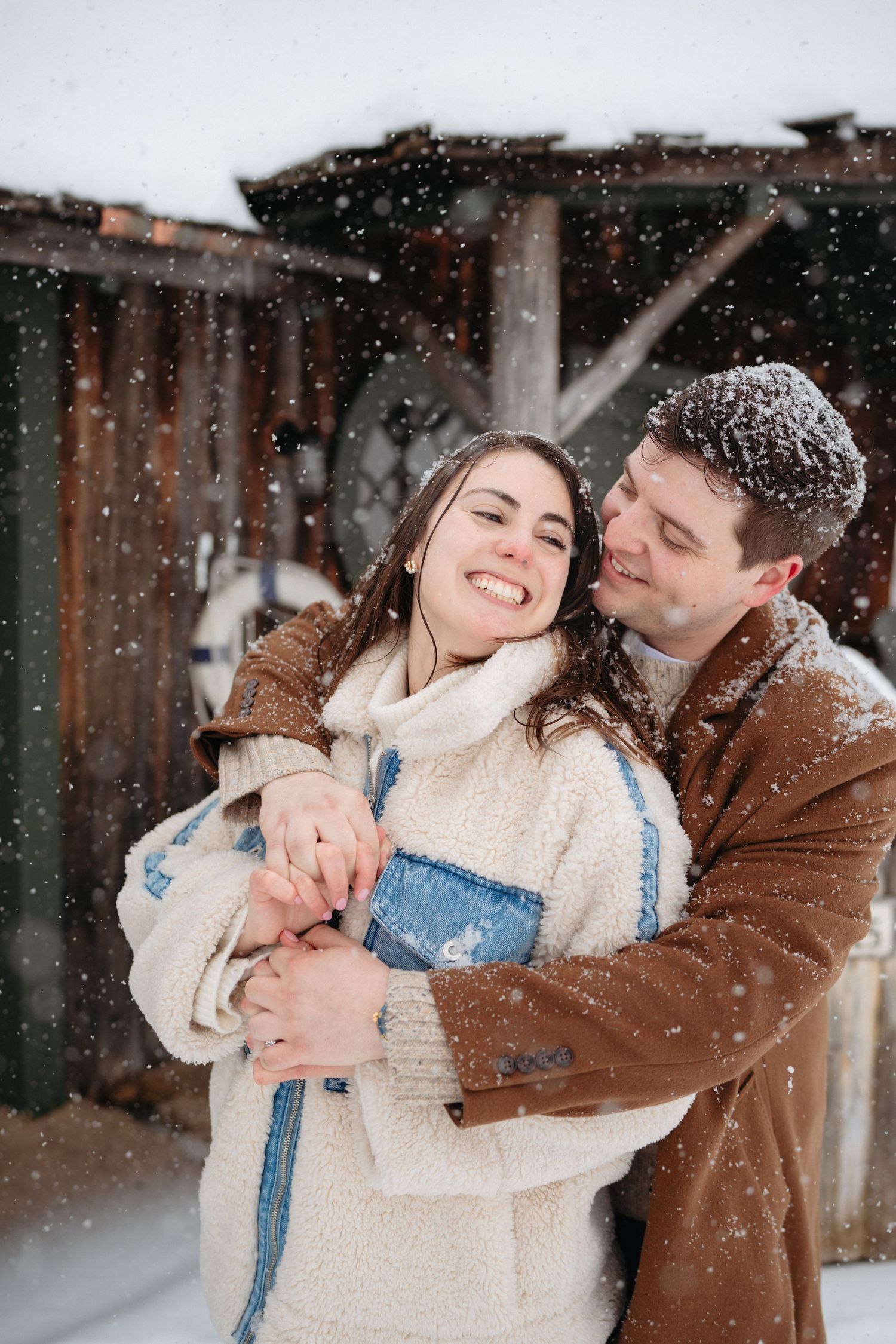 Happy couple embracing in snowfall wearing cozy winter coats outside a rustic wooden building.