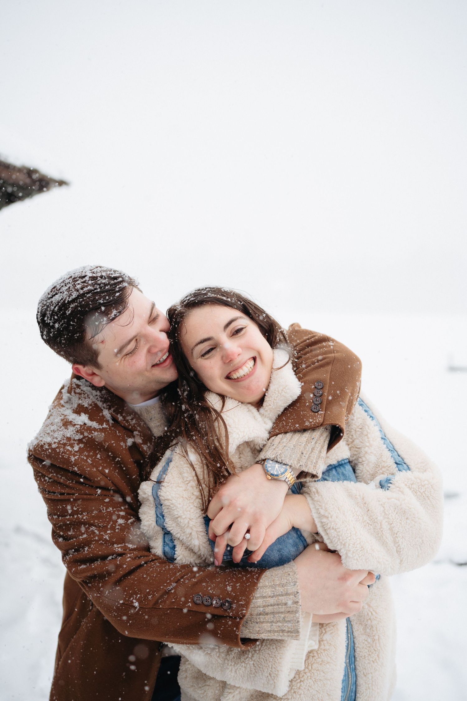 Happy couple hugging and laughing in heavy snowfall, man in brown coat embracing woman in white fluffy jacket outdoors.