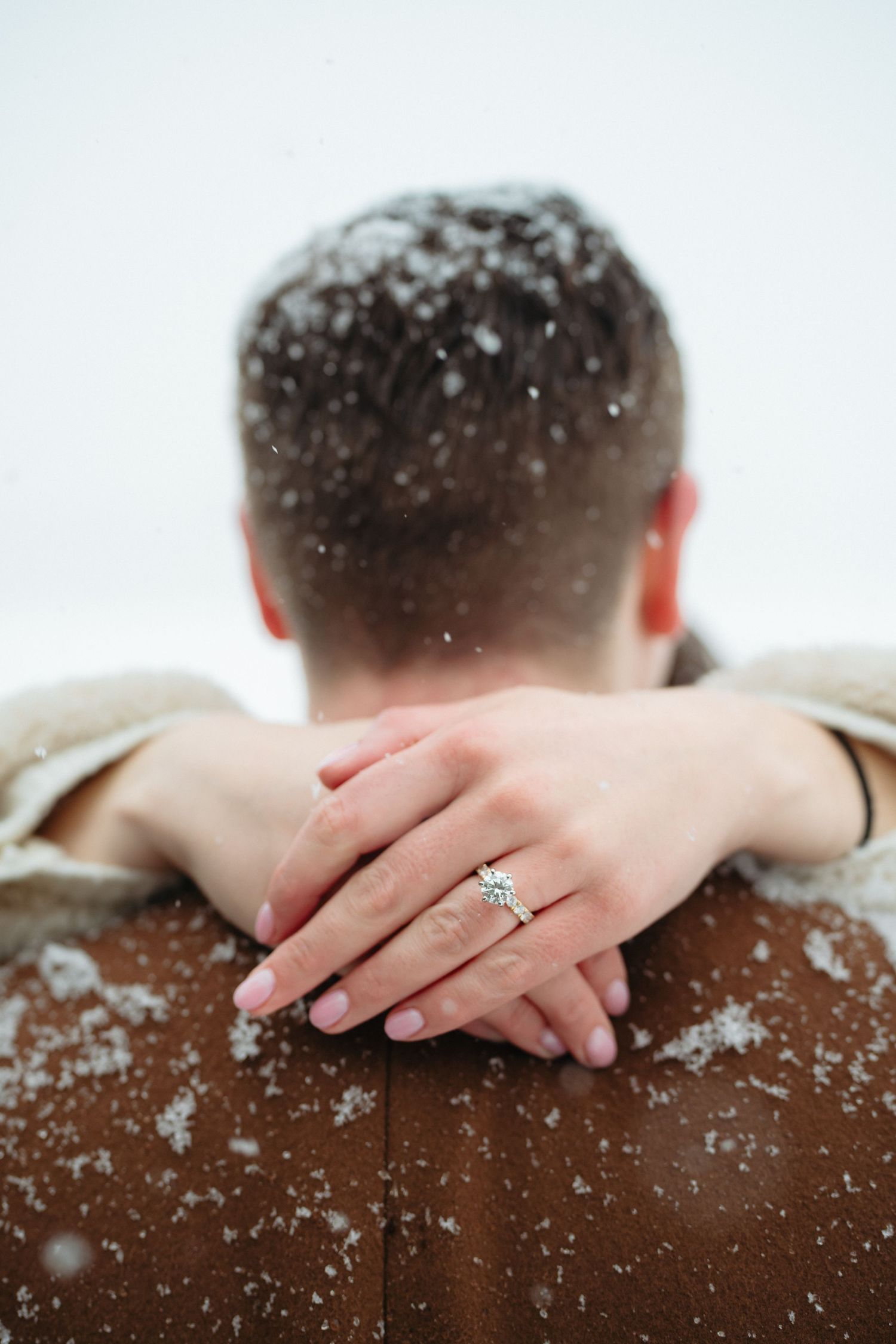 Close-up of womans hands with diamond engagement ring resting on snow-dusted brown jacket shoulders of her fiance.