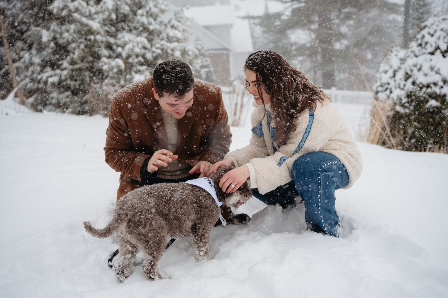 Couple crouching in snow with their dog during a winter snowstorm, wearing cozy jackets outdoors.