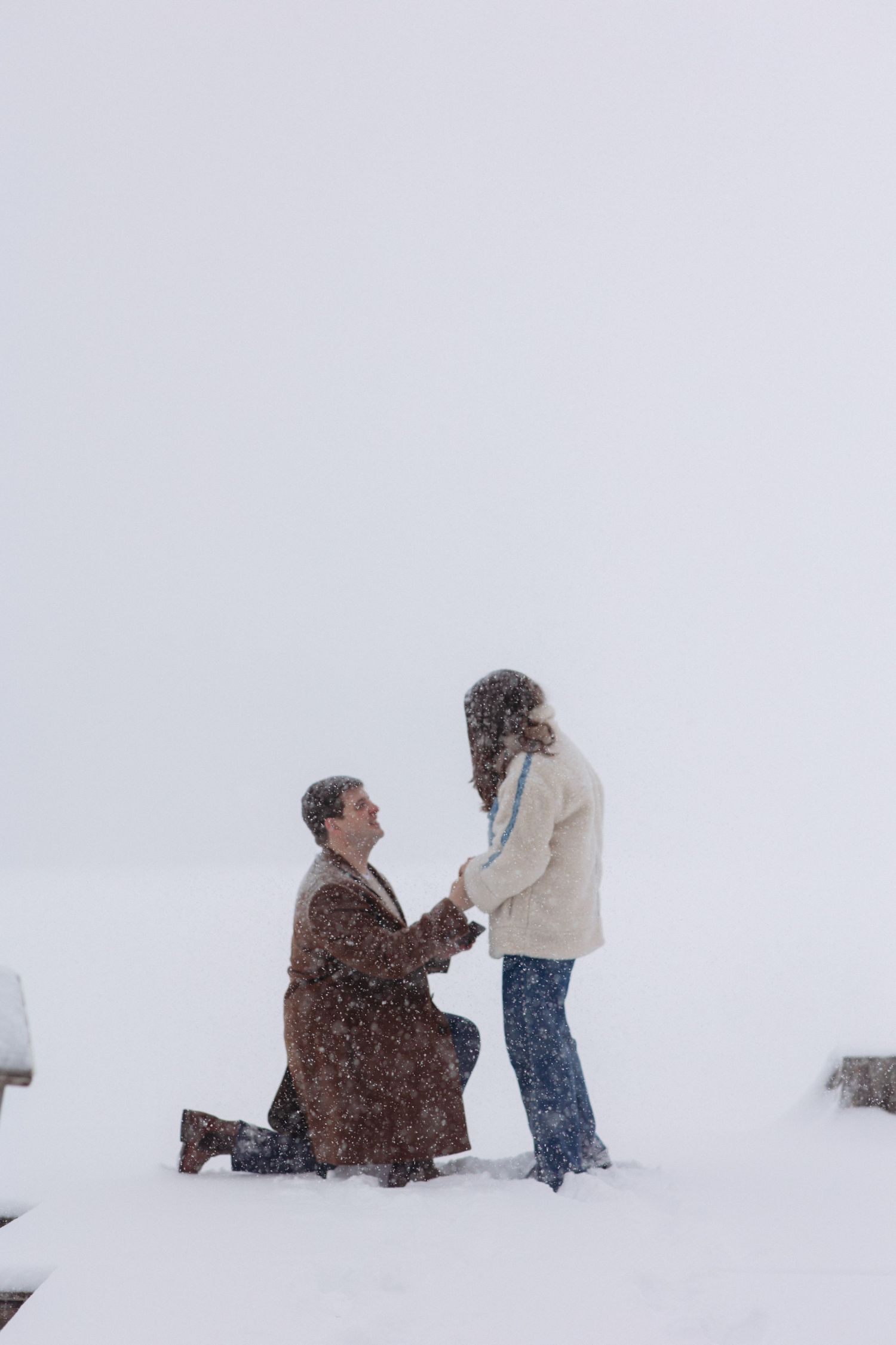 Man proposes on one knee to woman in snowy winter landscape, both wearing coats during a romantic snowfall.