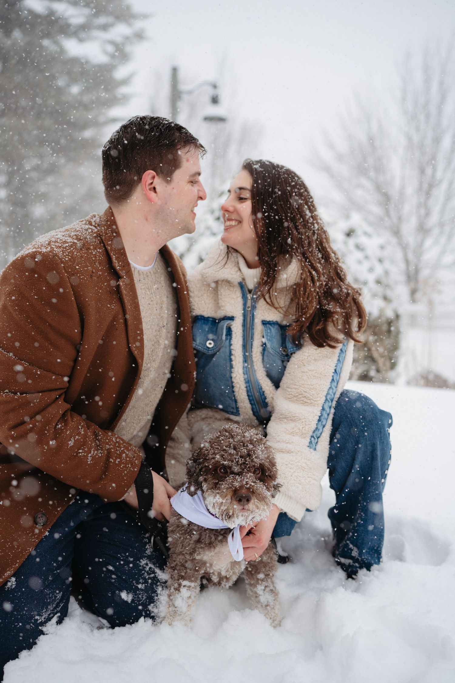 Smiling couple in winter jackets holding their curly brown dog while snow falls around them outdoors.