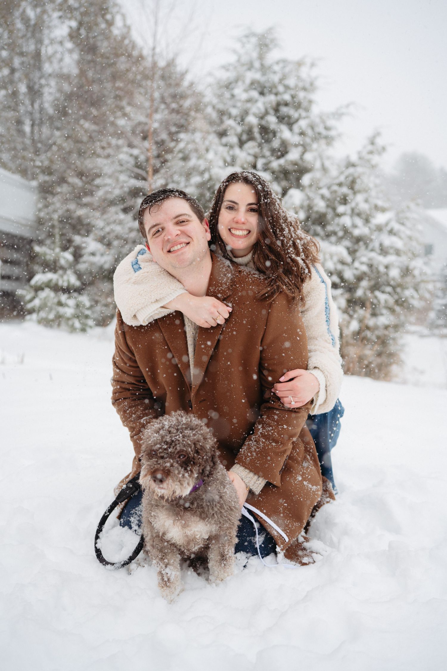 Happy couple with curly-haired dog posing in heavy snowfall, surrounded by snow-covered evergreen trees in winter.
