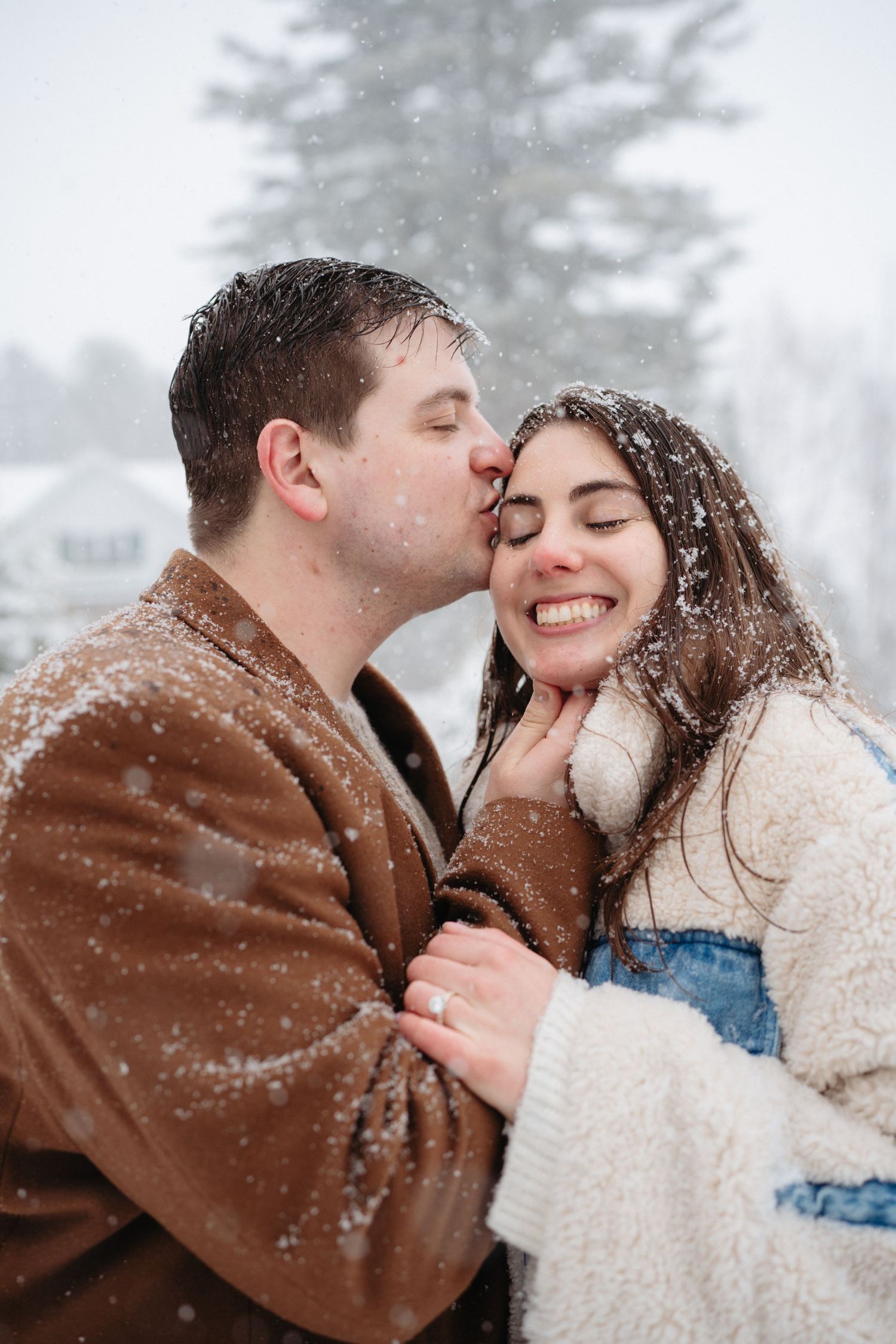 Man in brown coat kissing smiling womans cheek in snowy winter setting, both covered in fresh falling snowflakes.