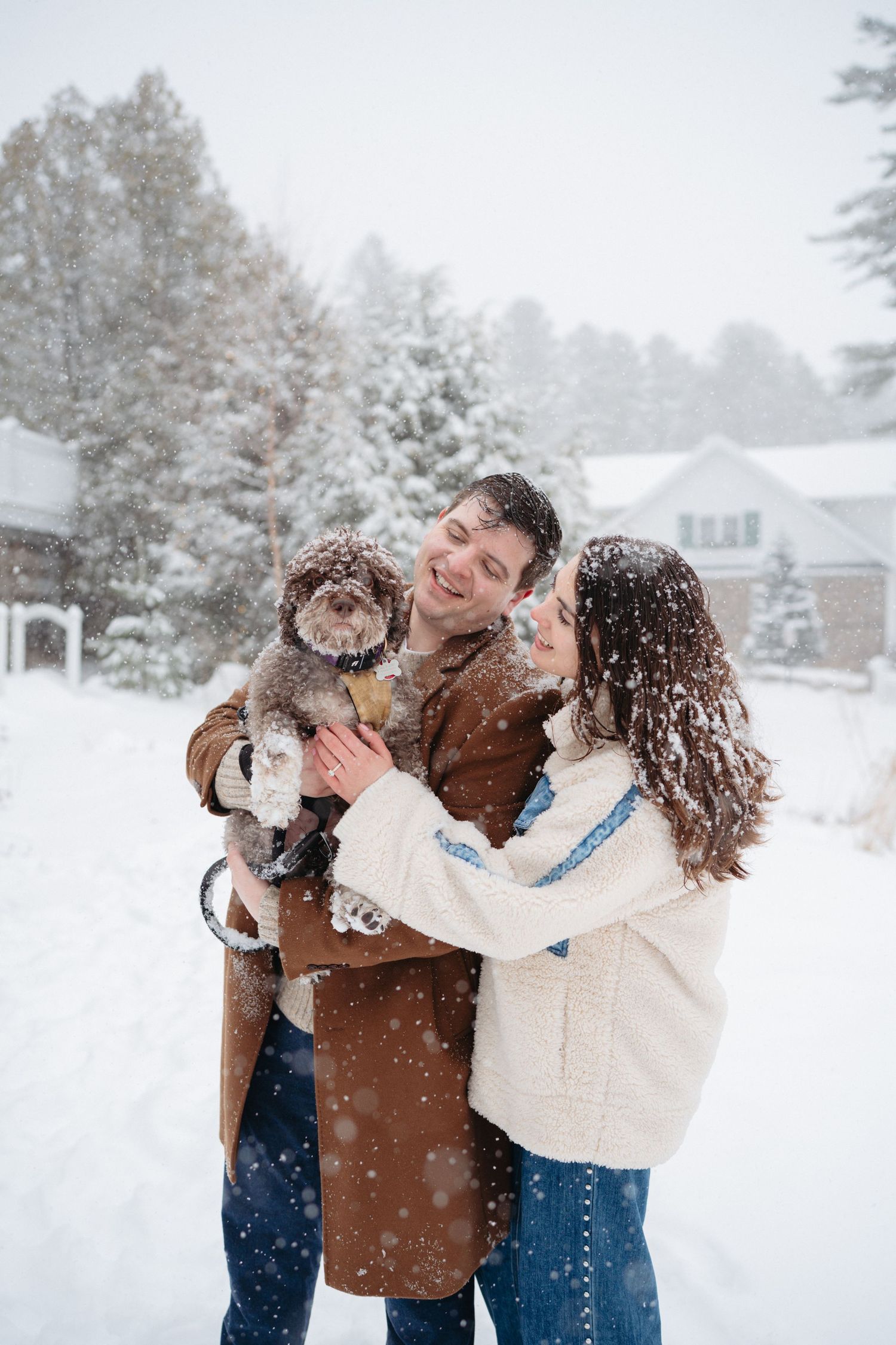 Couple smiling and holding a small dog in a snowy winter yard with snow-covered trees and a house in the background.