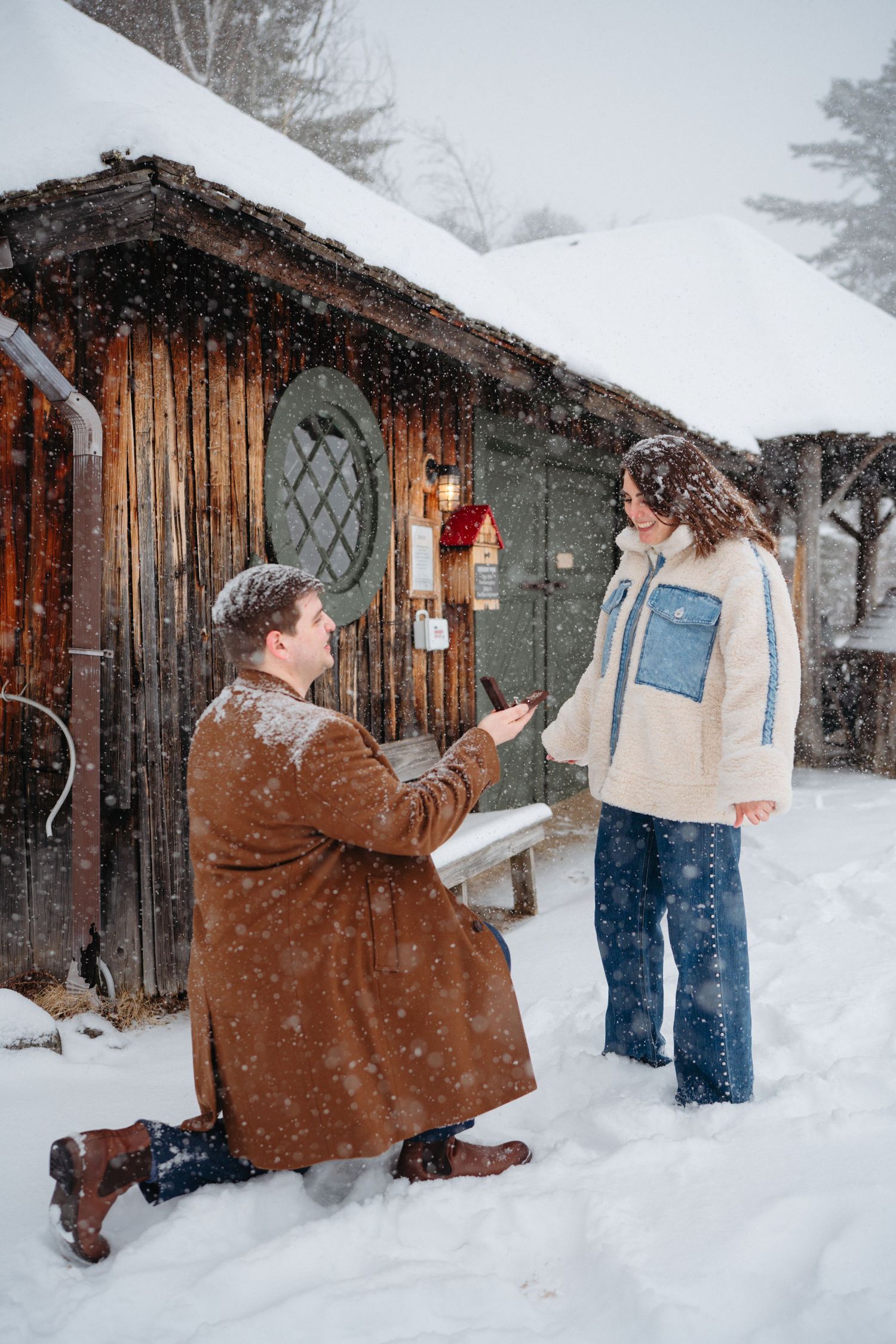 Man proposes on one knee in snowy winter setting outside rustic wooden cabin, woman reacts with surprise.