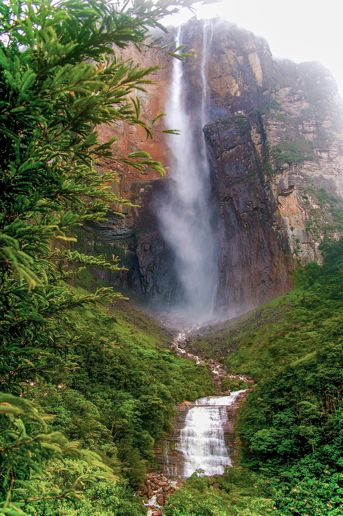 Towering waterfall cascades down steep rocky cliff face surrounded by lush green vegetation.
