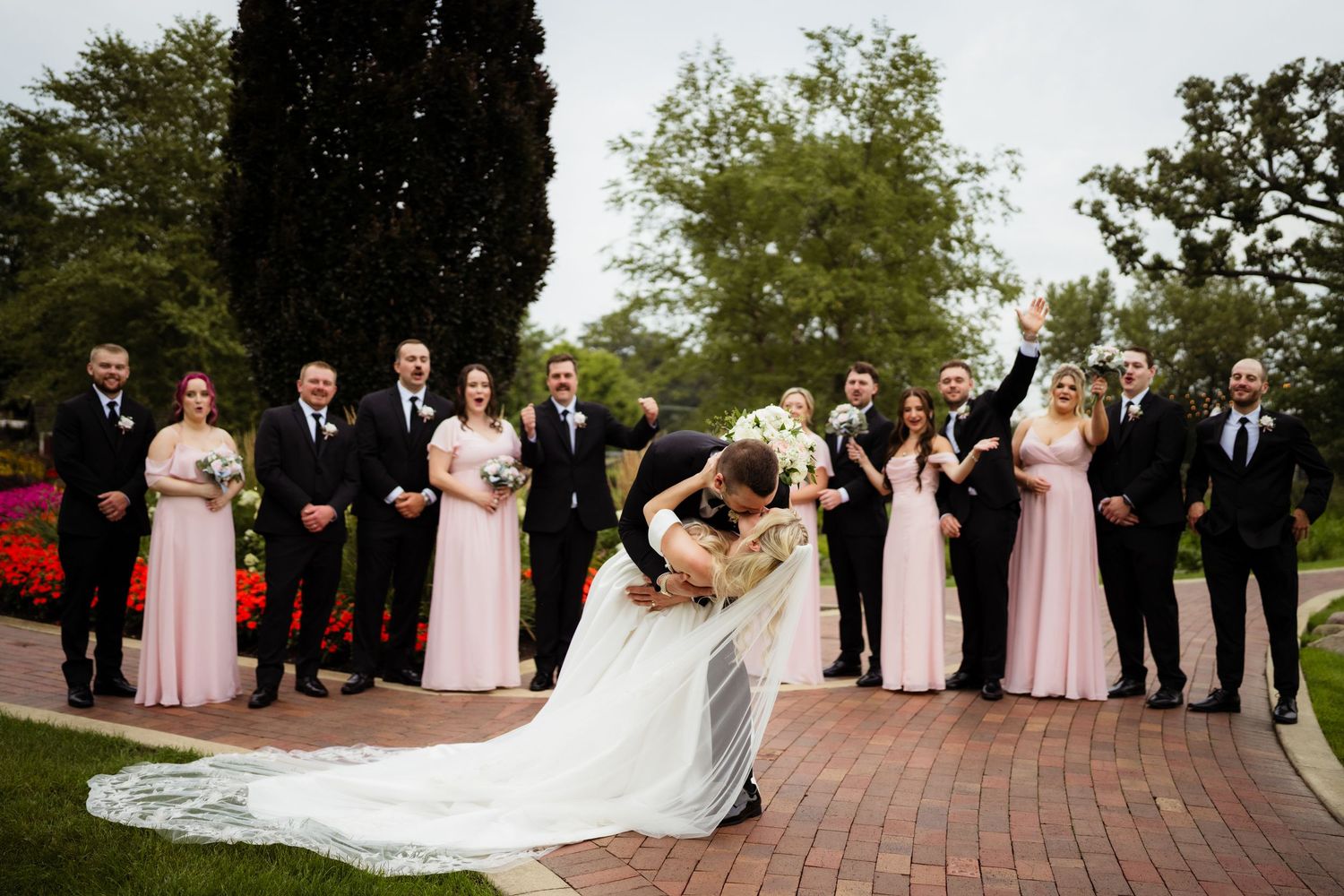 Bride and groom share a romantic dip kiss surrounded by wedding party in pink gowns and black suits in a garden.