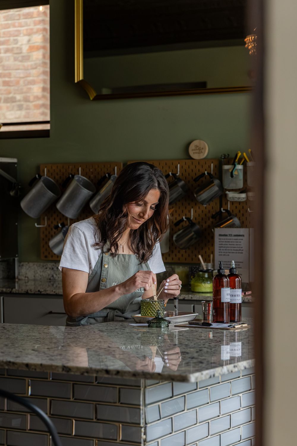A woman in a green apron prepares drinks at a stylish cafe counter with hanging mugs and green walls.