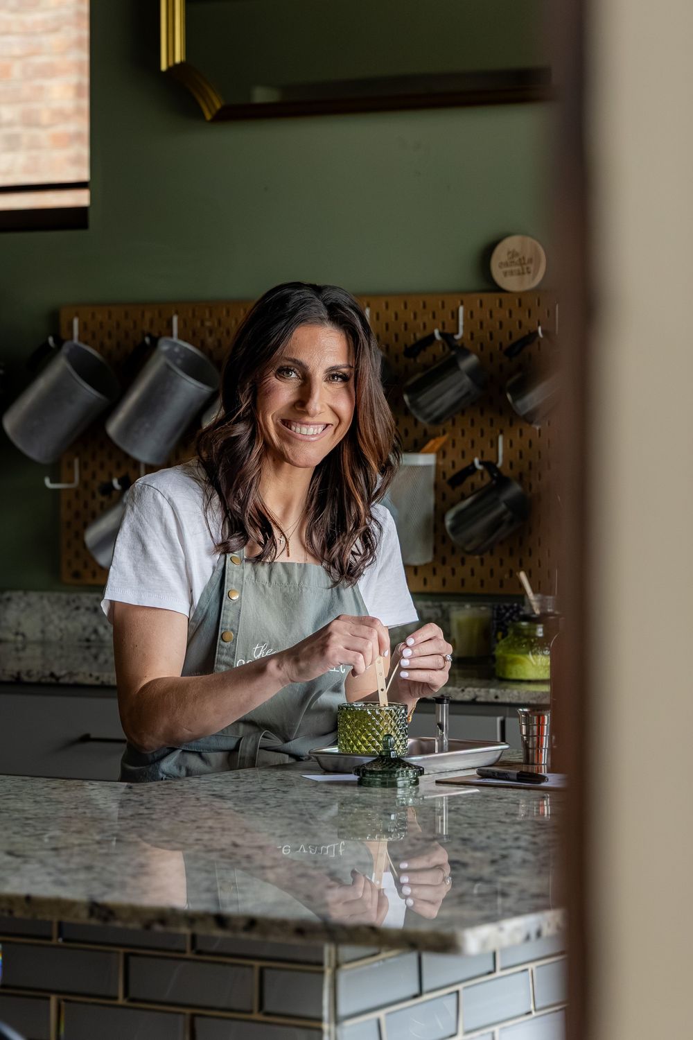 Smiling woman in green apron working at a marble kitchen counter with dark mugs hanging on a pegboard wall.