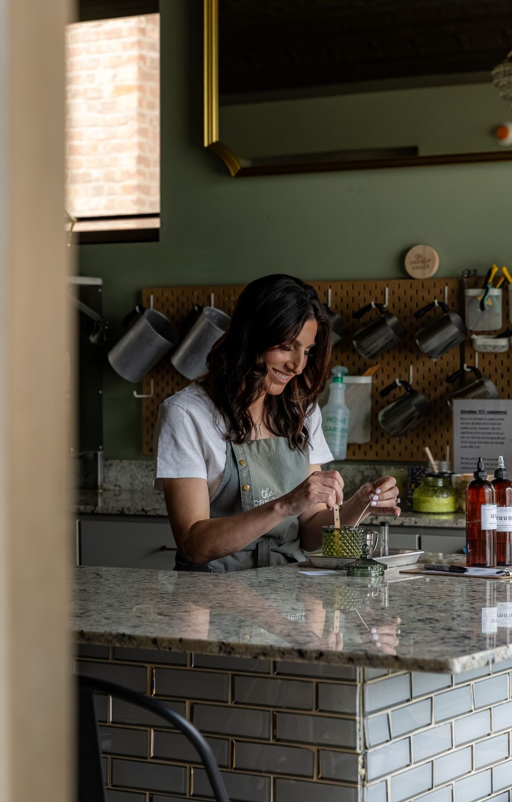 A smiling woman in an apron prepares food at a marble kitchen counter with hanging pots and green walls.