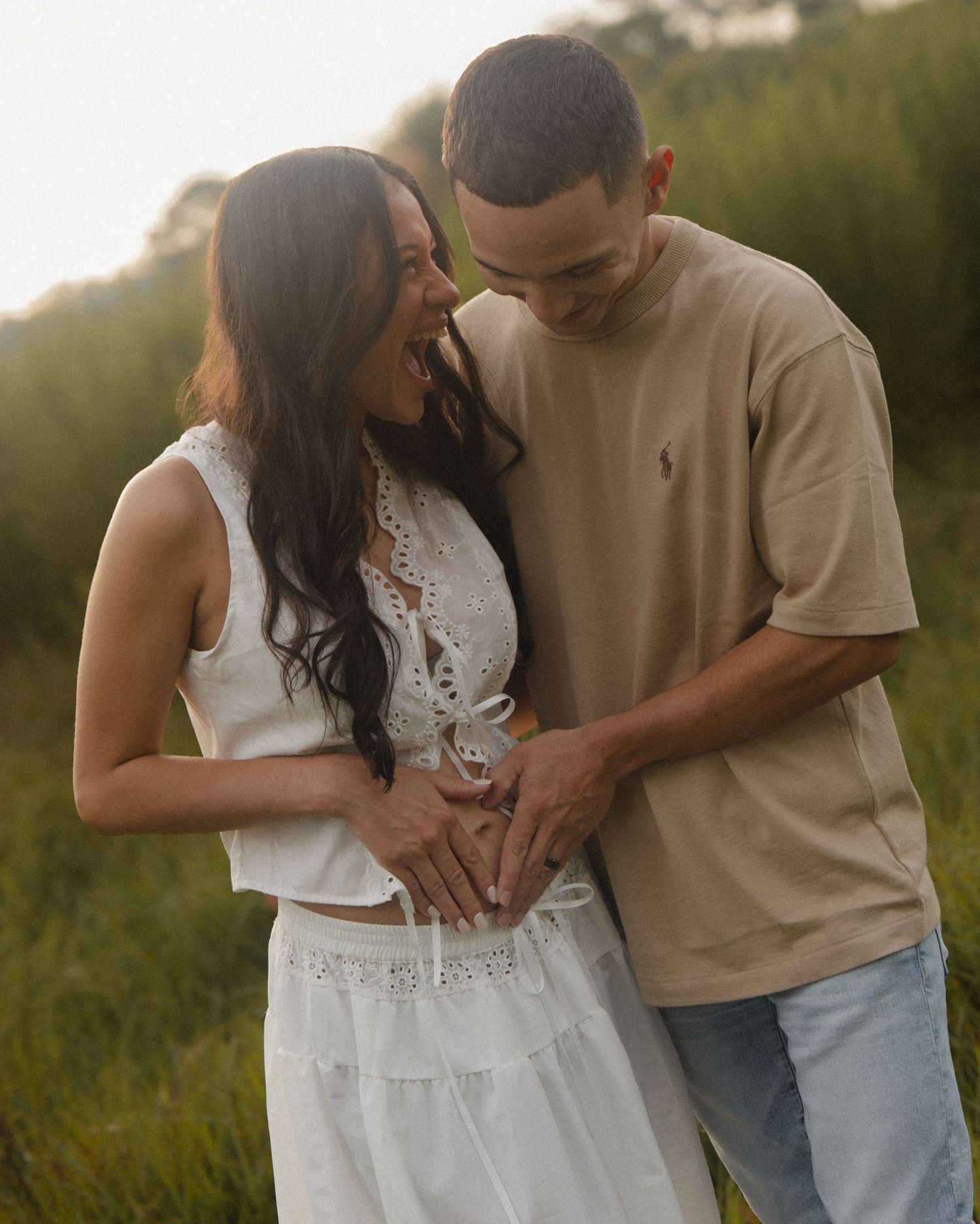 Happy couple laughing and cradling a baby bump together in a sunny green field.