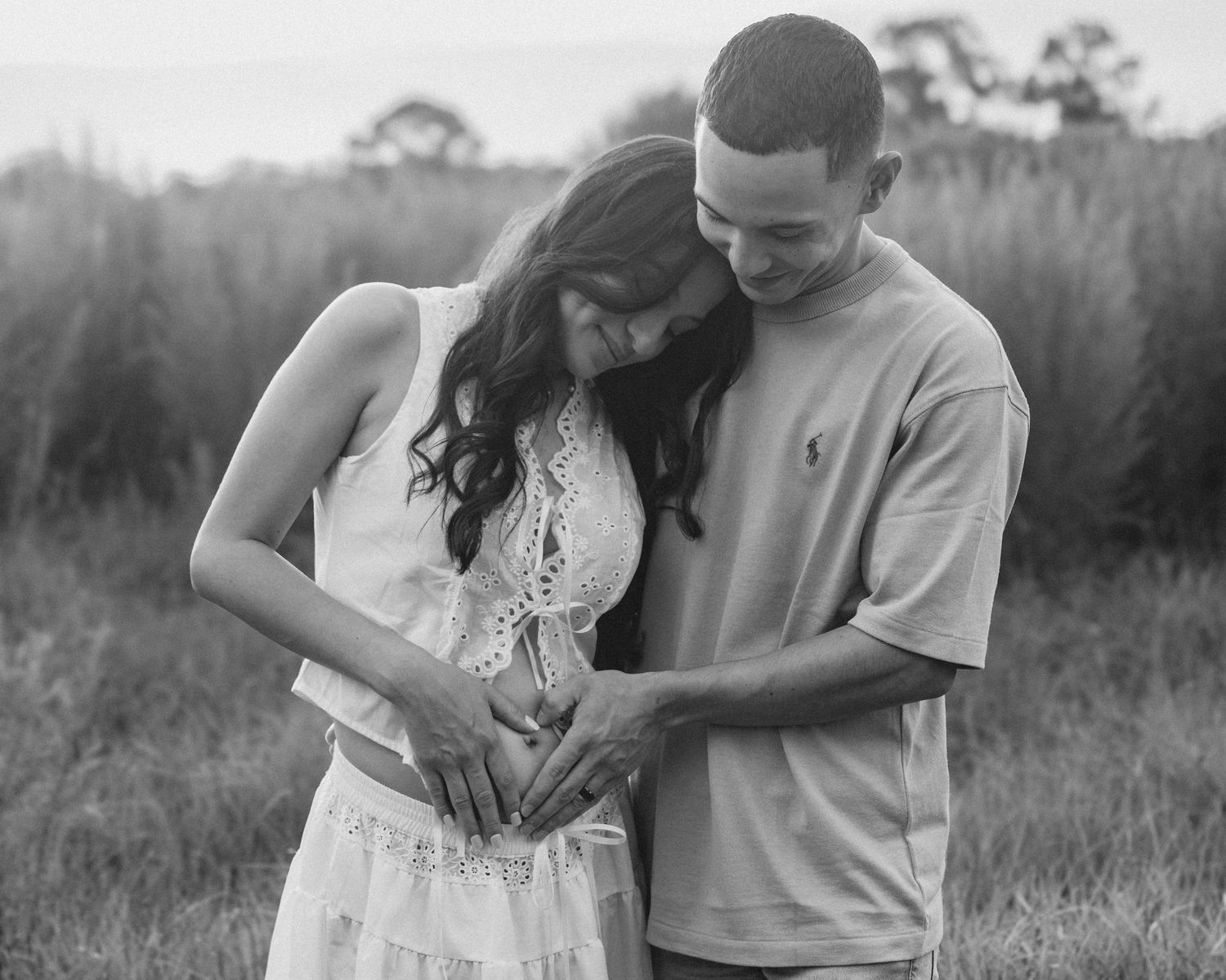 Black and white maternity photo of couple looking down at pregnant belly forming heart with hands.