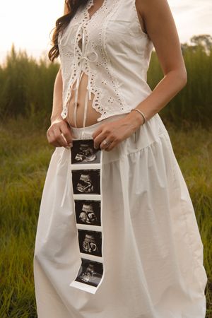 Pregnant woman in a white dress holding ultrasound photos in a lush green outdoor setting.