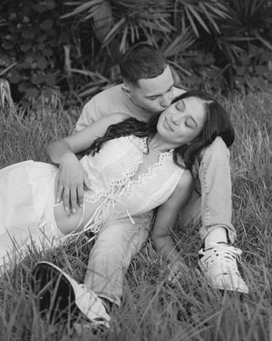 Black and white photo of a couple sitting in grass, man kissing woman's cheek during maternity photoshoot.