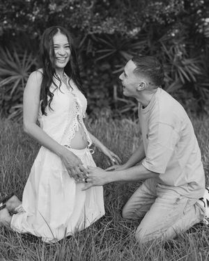 Black and white maternity photo of smiling pregnant woman in white dress with partner kneeling in grass.