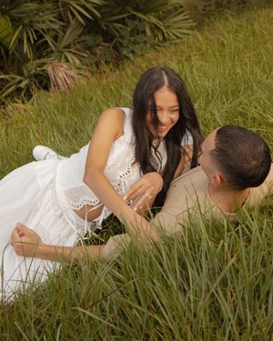 Couple sharing a joyful moment lying in tall green grass during outdoor maternity photoshoot.
