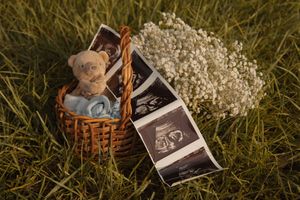 Wicker basket with teddy bear, ultrasound photos, and baby booties with white flowers in grass.