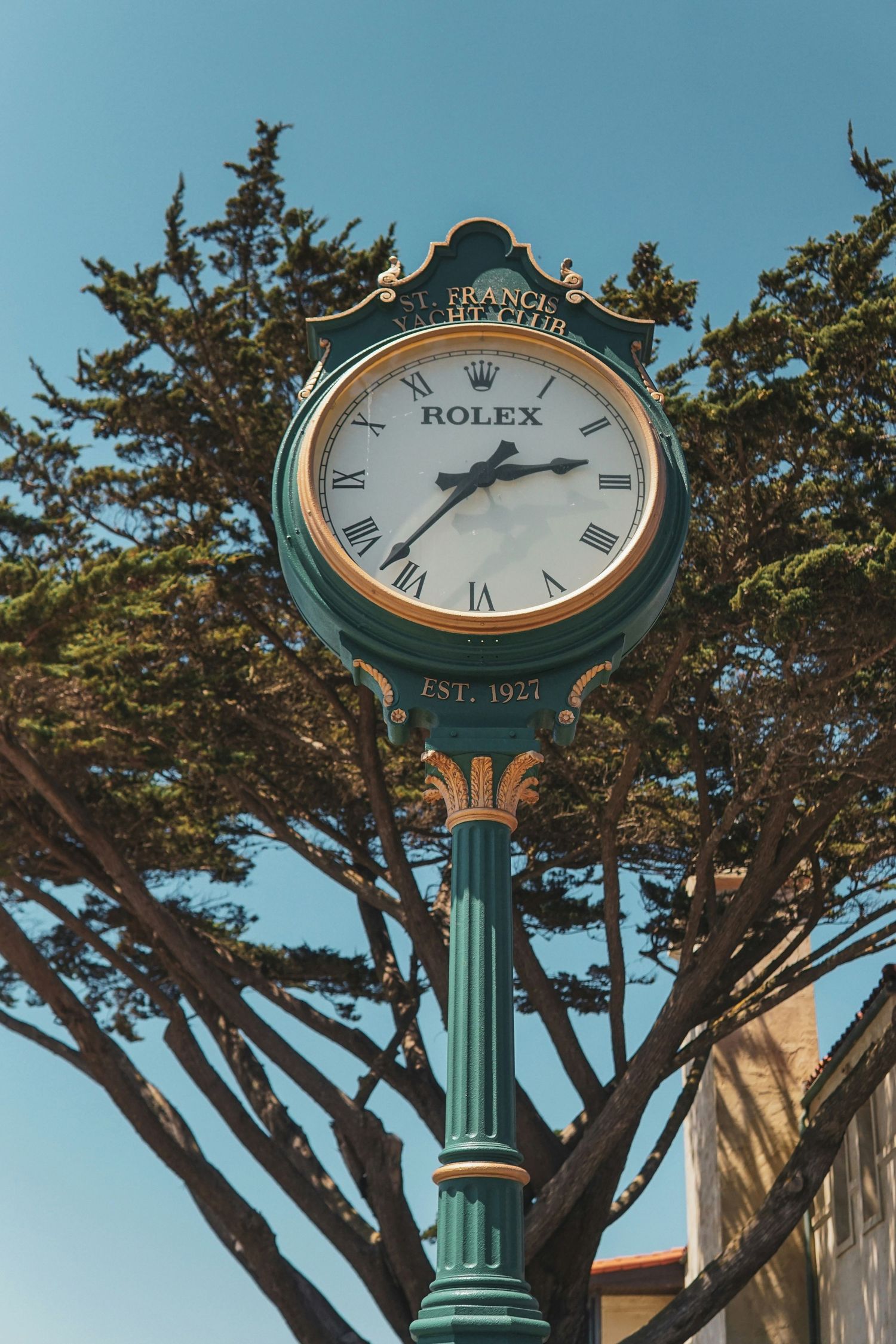Elegant green Rolex street clock with Roman numerals on ornate post, EST. 1927, against blue sky and trees.