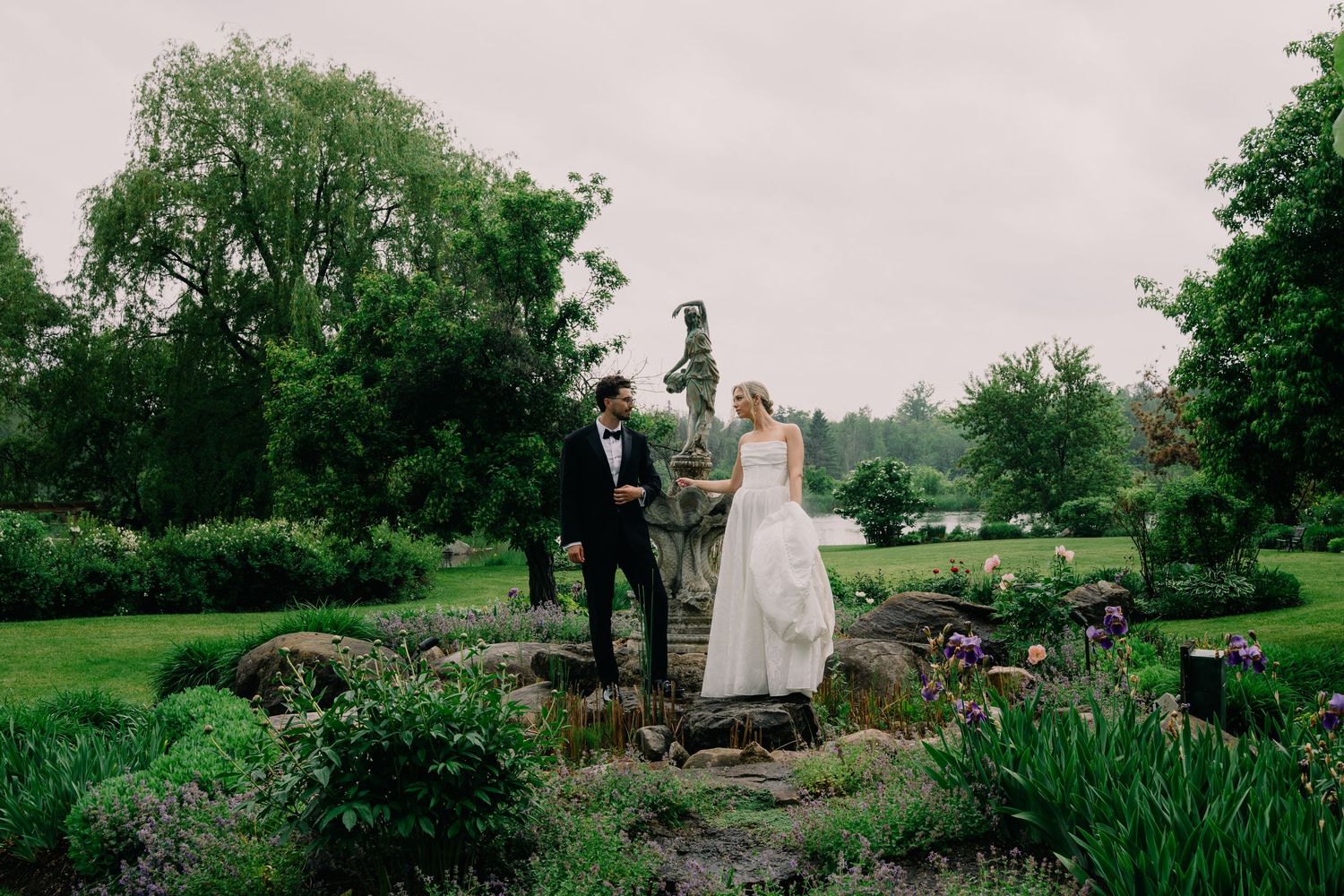 Bride and groom stand together in a lush garden with stone bridge, trees, and flowers on their wedding day.