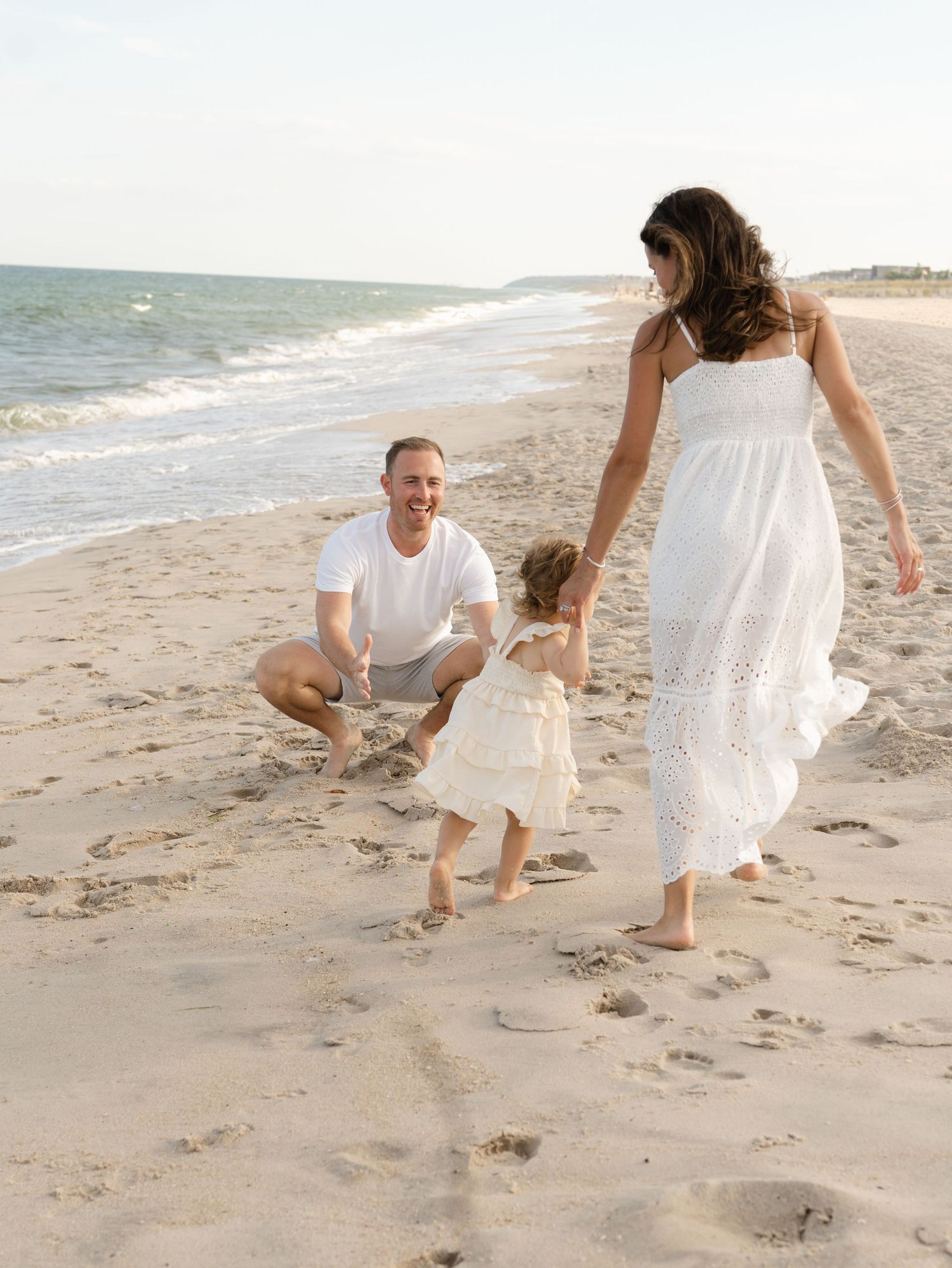 Toddler girl runs to her father along the shoreline while her mother holds her hand.
