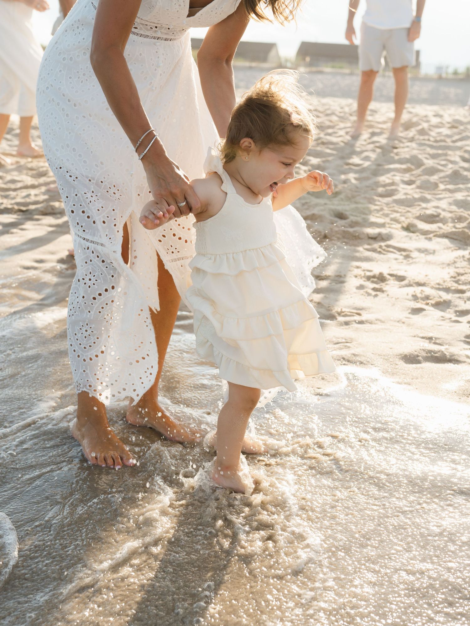 A mother in a white dress holds her daughter as she jumps in the ocean waves near Jenkinsons.