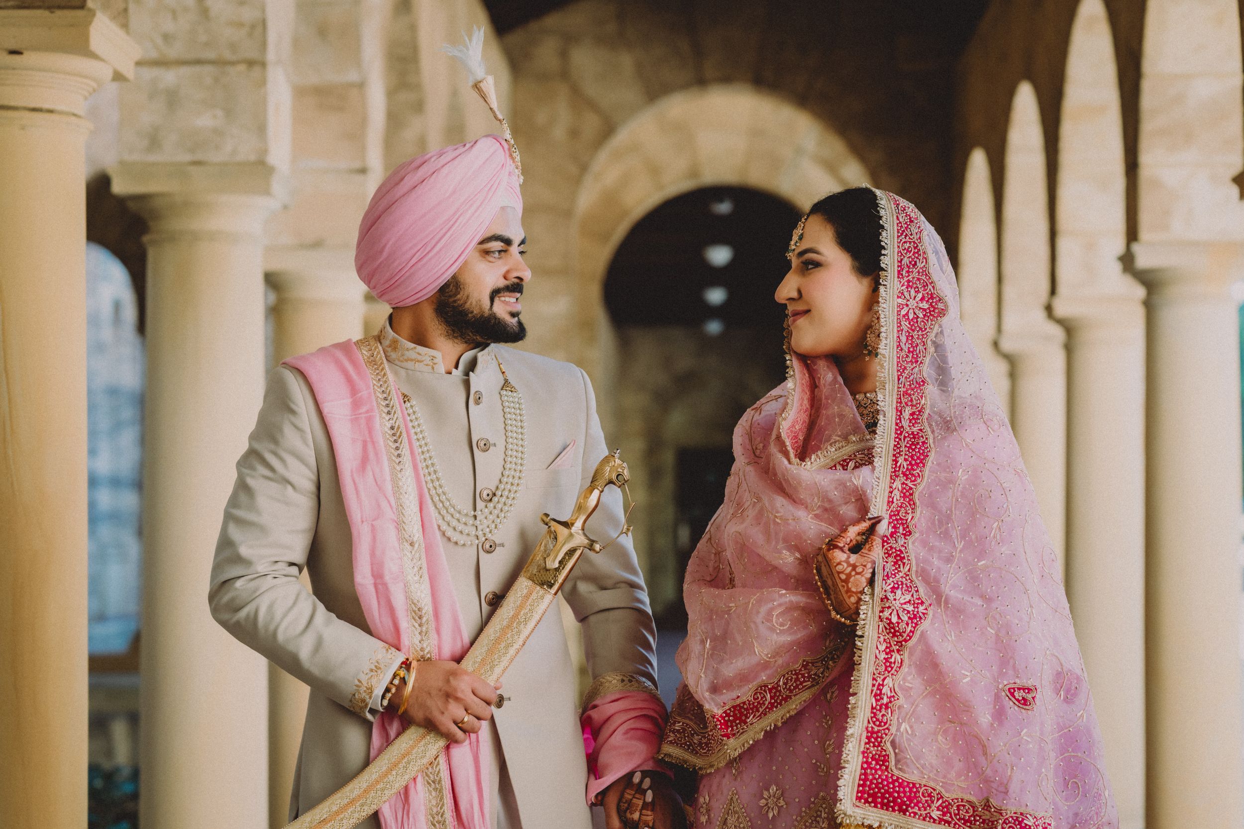 Sikh couple in traditional wedding attire, groom in gold sherwani with pink turban, bride in pink lehenga with dupatta.