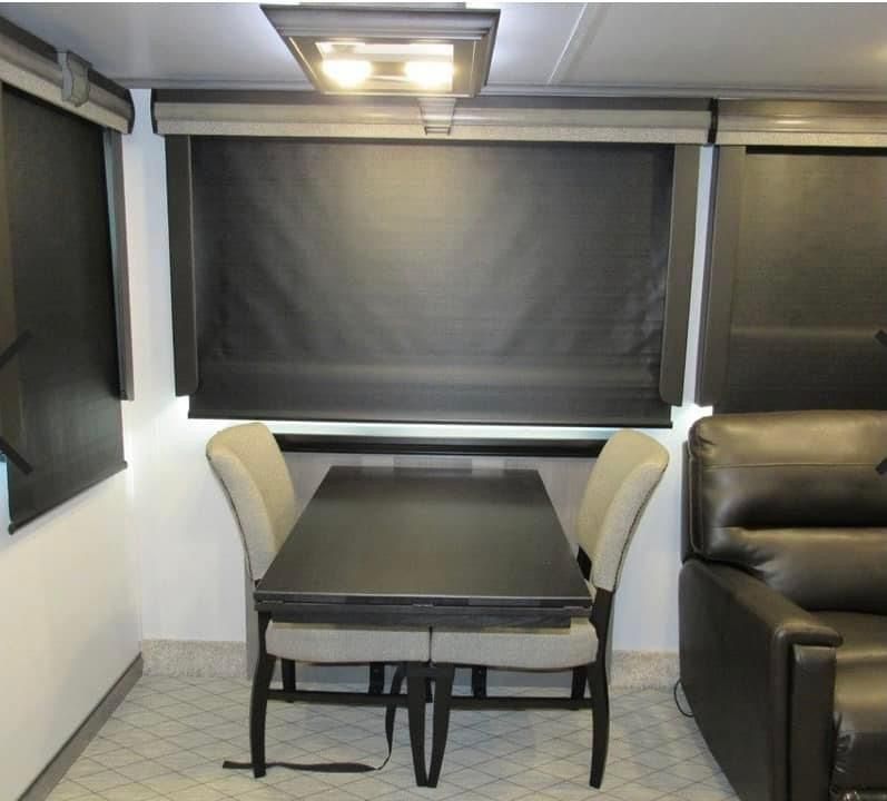 RV dining area with dark table, two chairs, blackout roller shades, and leather sofa.