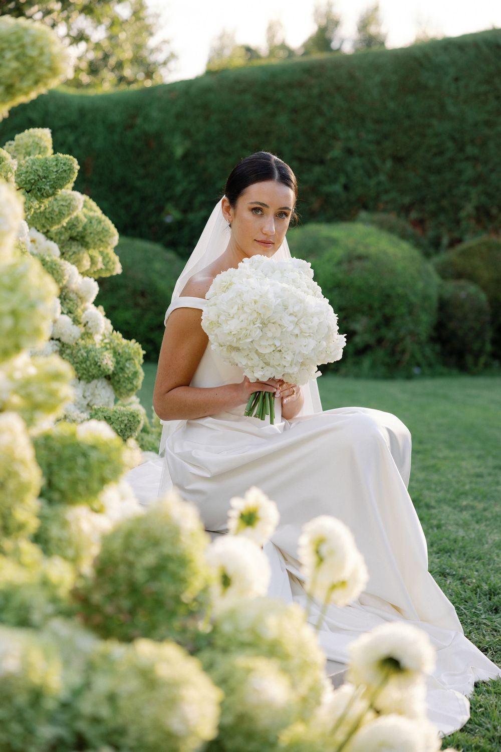 Bride in white gown holding hydrangea bouquet seated among lush green garden florals wearing veil.